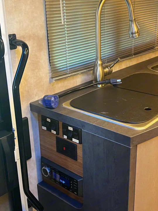 Inside an RV rental: a kitchen counter with a modern sink, faucet, and an unplugged power cord. Control switches and a stereo are mounted below.. Winnebago View 2019