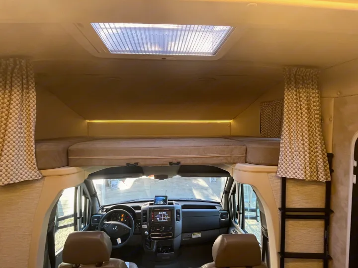 Spacious RV rental interior featuring a cab-over bunk bed, ceiling skylight, and front cockpit with dashboard & steering wheel.. Winnebago View 2019