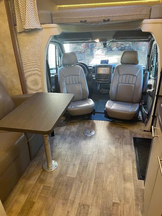 View into an RV rental's living area and cockpit. Features wood-look flooring, tan sofa, gray seats, table, and overhead bunk.. Winnebago View 2019