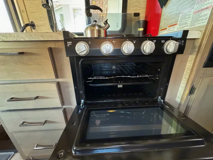 Inside an **rv rental** kitchen, a black gas stove and open oven with racks are visible. A kettle sits on the stovetop, with light-colored drawers to the left.. Thor Motor Coach Coleman 2021