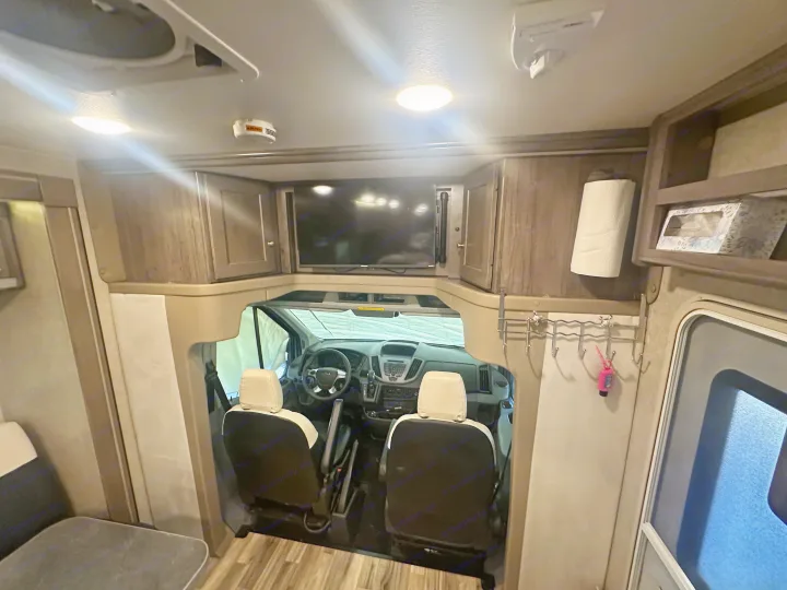 Interior of an RV rental showing the living area leading to the driver's cab with a mounted TV, seats, and overhead storage.. Winnebago Fuse 2017