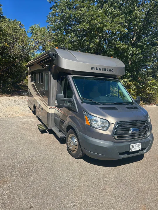 Sleek grey Winnebago RV rental, a Ford motorhome, parked outdoors under a sunny blue sky, surrounded by green trees.. Winnebago Fuse 2017