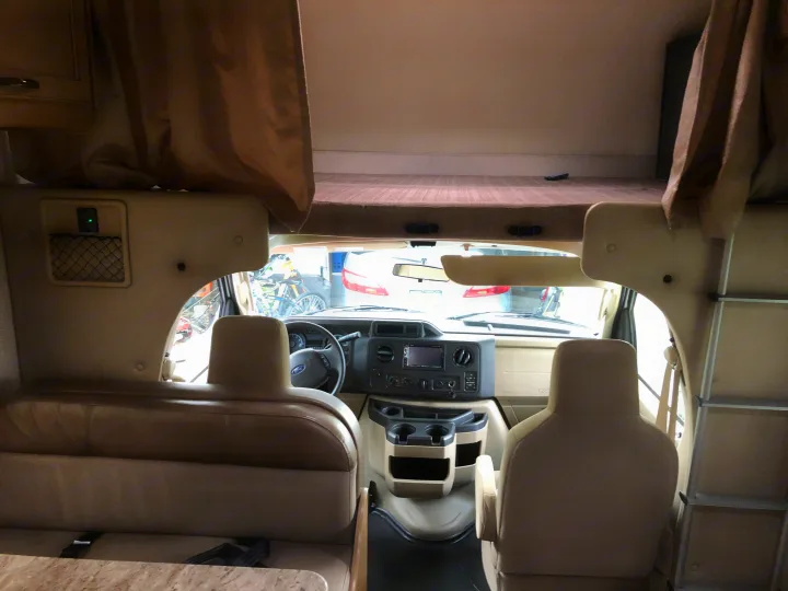 Interior of an RV rental: light beige cockpit with Ford steering wheel, dash screen, and a large overhead bunk accessible by ladder.. Thor Daybreak 2020