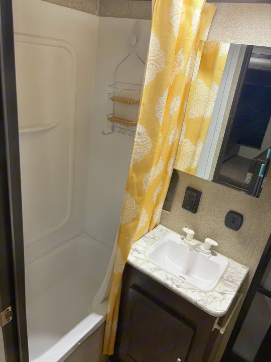 Inside an RV rental bathroom, featuring a white shower stall with a yellow patterned curtain and a sink with a marble-look counter.. Keystone RV Summerland 2018