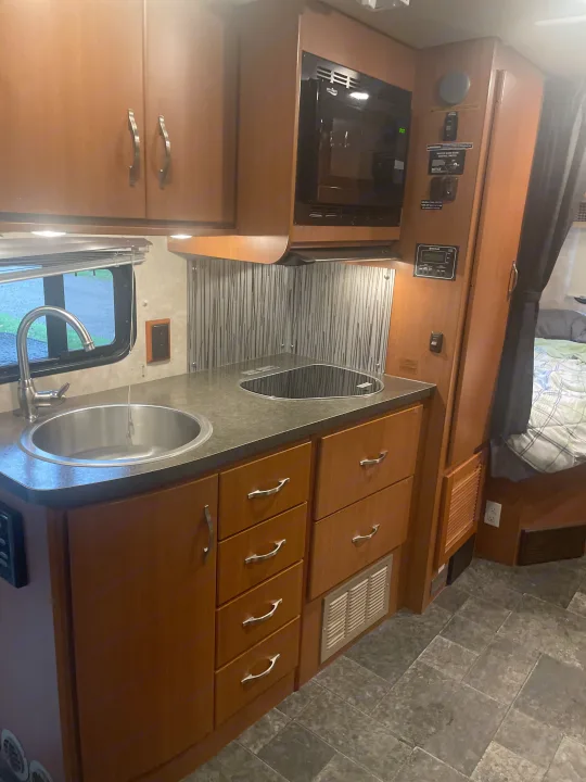 This RV rental's practical kitchen includes a stainless steel sink, black cooktop, microwave, and warm wood cabinetry.. Itasca Navion 2012