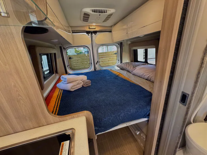 Inside an RV rental: a cozy bedroom with a blue blanket, folded towels, and rear windows, plus a compact kitchen setup.. Thor Tellaro 2022