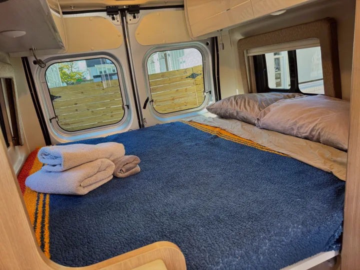 Cozy RV rental interior with a bed featuring a blue blanket, pillows, and folded towels. Rear windows reveal an outdoor fence and buildings.. Thor Tellaro 2022