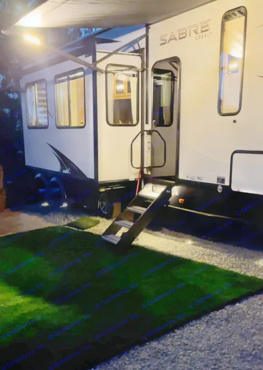 A white SABRE COBALT RV rental illuminated at night, showing an extended awning, glowing windows, and steps on gravel.. Forest River Sabre 2023