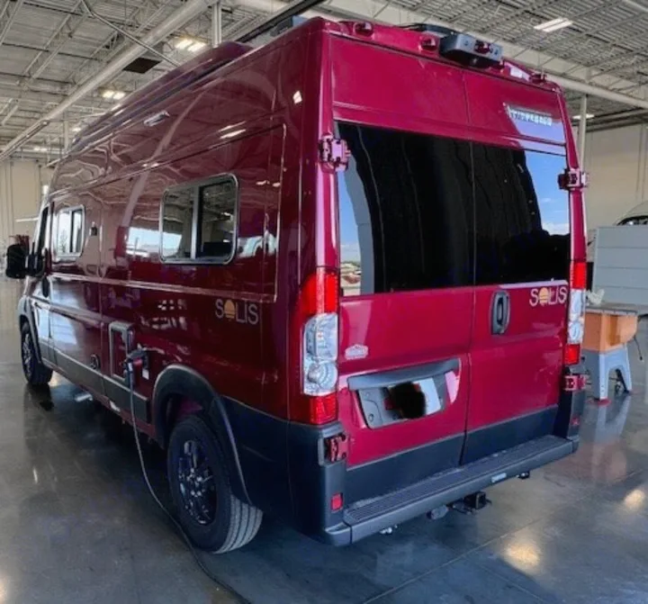 This deep red Winnebago Solis camper van, ideal for rv rental, is seen from the rear 3/4 view while parked indoors.. Winnebago Solis 2023