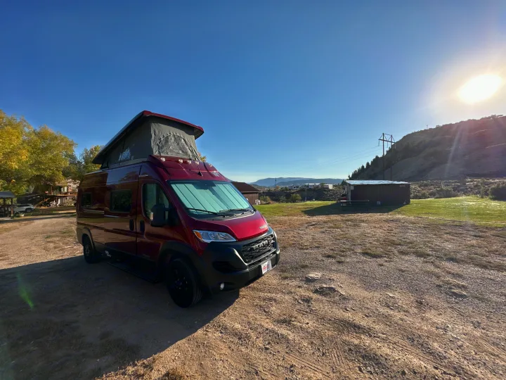 A vibrant red pop-top camper van, perfect for an RV rental, parked on a sunny dirt lot with hills & buildings.. Winnebago Solis 2023