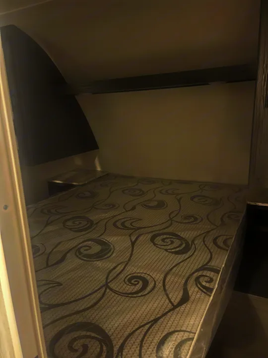 RV rental bedroom features a comfy bed with a new, plastic-covered mattress, built-in nightstands, and overhead shelf.. Dutchmen 26bhc 2024