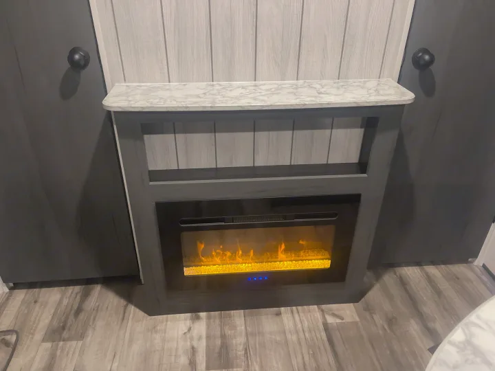 A built-in electric fireplace with glowing faux flames, a marble-look counter, and wood paneling in a modern RV rental.. Dutchmen 26bhc 2024