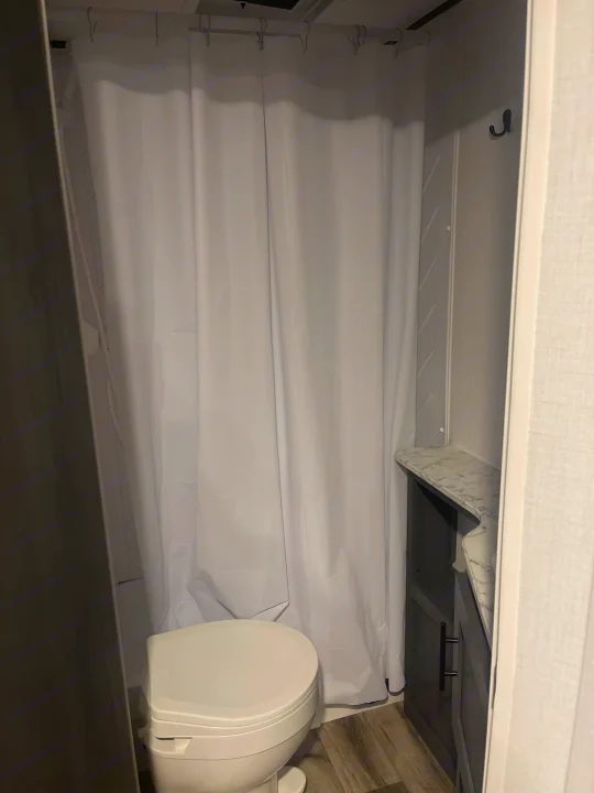 A compact RV rental bathroom featuring a toilet, white shower curtain, and small vanity with a sink.. Dutchmen 26bhc 2024