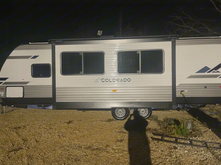 Side view of a white & grey Colorado travel trailer, parked on gravel under artificial light at night. Consider for an RV rental.. Dutchmen 26bhc 2024