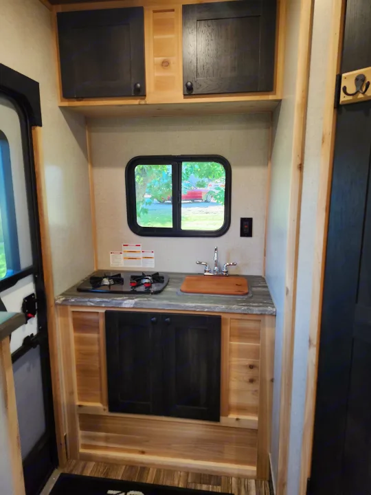 Compact RV rental interior featuring a kitchenette with a two-burner stove, sink, and black/wood cabinets, plus a window.. Glacier Ice House 17RBLE 2023