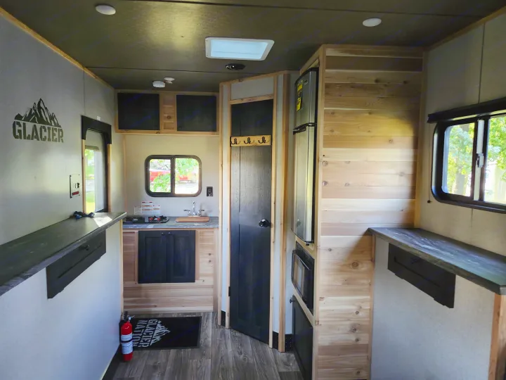 Inside this RV rental: a compact kitchen with stove, sink, refrigerator, and microwave, plus cozy wood paneling.. Glacier Ice House 17RBLE 2023
