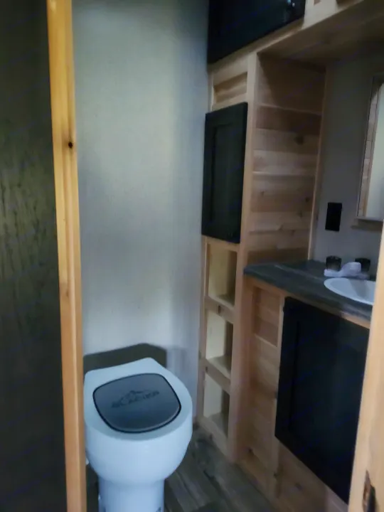 This compact RV rental bathroom features a white toilet, sink on a black counter, and warm wood shelving and cabinets.. Glacier Ice House 17RBLE 2023