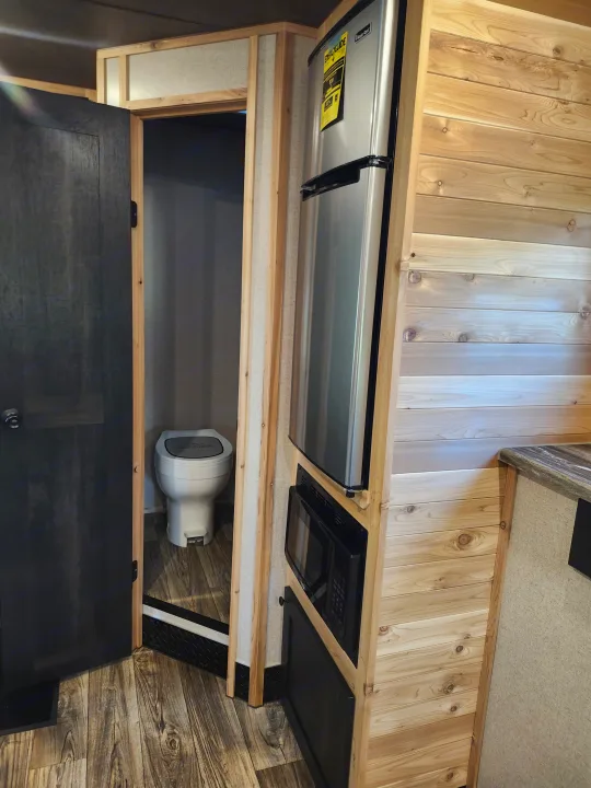 RV rental interior: compact kitchen with fridge/microwave, and a small bathroom with toilet visible. Wood-paneled walls.. Glacier Ice House 17RBLE 2023