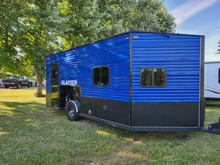 Bright blue & black Glacier ice house trailer, ideal for RV rental, parked on green grass with trees in the background.. Glacier Ice House 17RBLE 2023