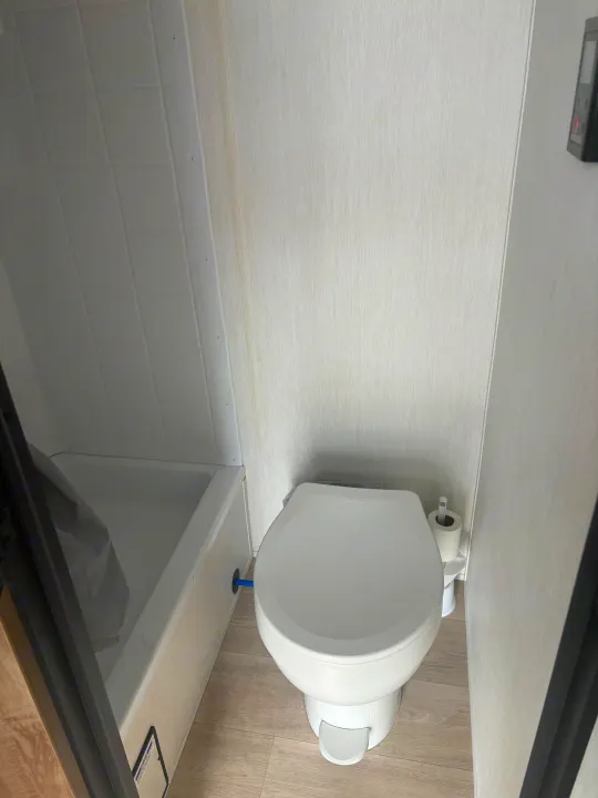 Compact RV rental bathroom featuring a white toilet with a foot pedal, a small shower/tub, and light textured walls.. Forest River Wolf Pup 2025