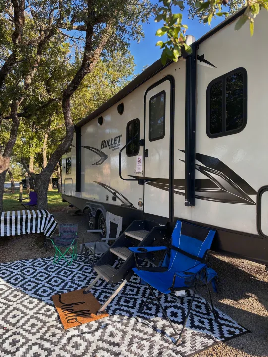 Enjoying the outdoors with this RV rental campsite, featuring a bright travel trailer, patterned mat, and seating under trees.. Keystone RV Bullet Ultra Lite 2021