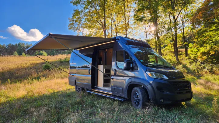 Enjoy nature with this RV rental! Dark grey camper van with awning extended in a sunny field.. Airstream Rangeline 2024