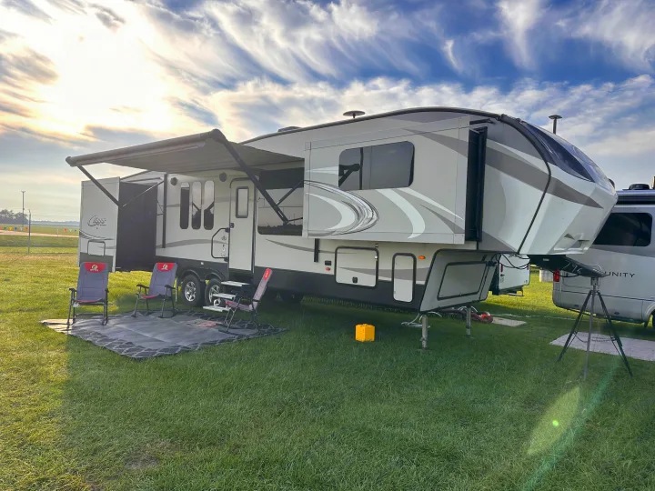 A large fifth-wheel RV rental is set up on a grassy campsite with an extended awning, two chairs, and mat, under a cloudy sky.. Keystone RV Cougar 2017