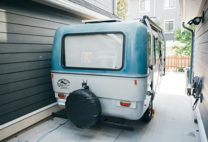Compact blue and white RV rental parked in a narrow driveway between buildings. Features a rear window and spare tire.. Outback Trailer Manufacturing the Outback 2017