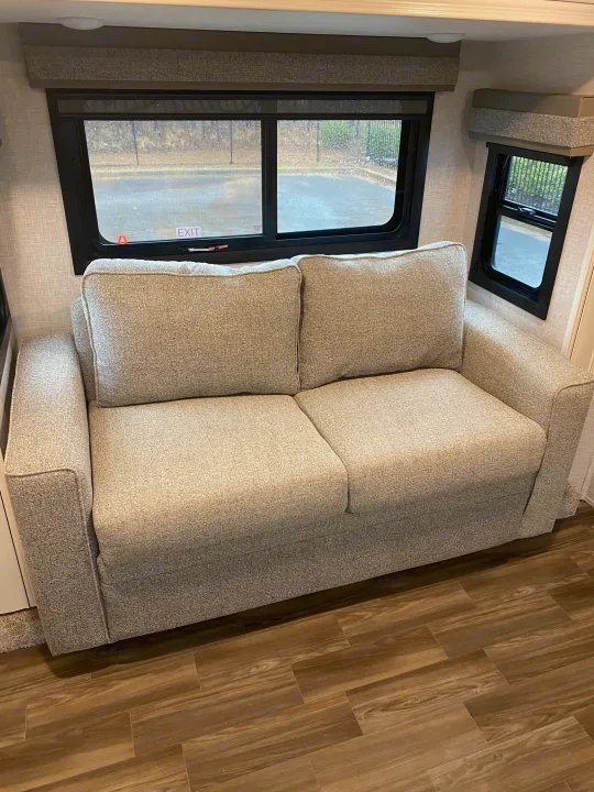 Light gray fabric sofa inside an RV rental, with a view through two windows to a wet exterior and wood-look flooring.. Highland Ridge RV Open Range 2024