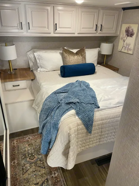Cozy RV rental bedroom with white bedding, blue accents, overhead cabinets, lamps, and a patterned rug.. Highland Ridge RV Open Range 2024