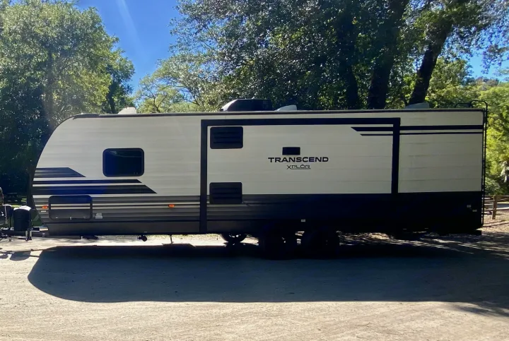 A white and dark gray Transcend XPLOR RV rental is parked on a sunny campsite, surrounded by green trees.. Grand Design Transcend Xplor 2021