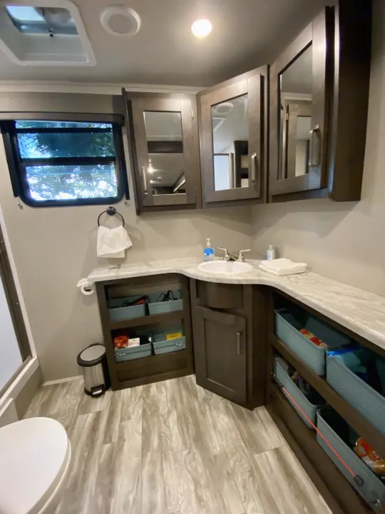 Modern RV rental bathroom featuring a well-lit vanity, sink, mirrored cabinets, and open-shelf storage with bins.. Grand Design Transcend Xplor 2021