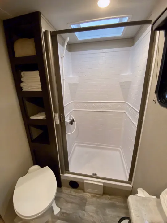 Compact RV rental bathroom: a clean shower stall with skylight, toilet on left, and towel storage shelves.. Grand Design Transcend Xplor 2021