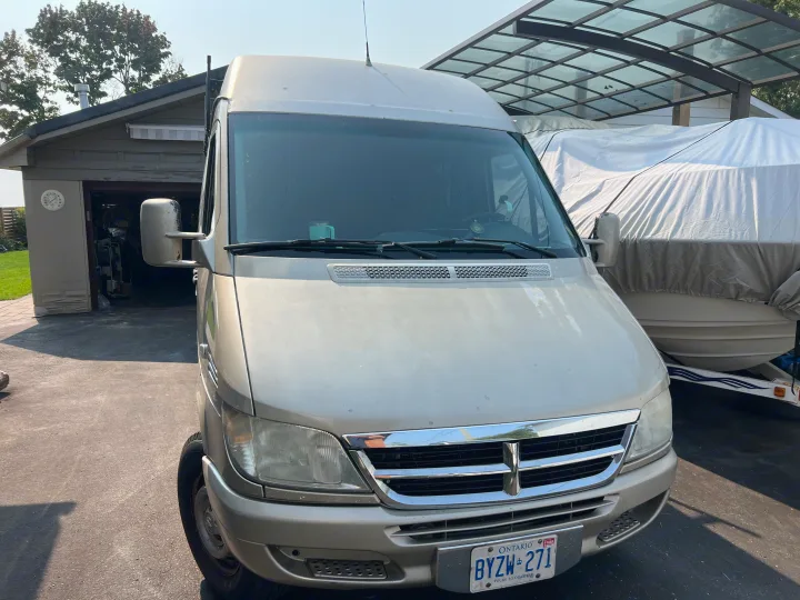 Light-colored camper van, an ideal rv rental, parked in a driveway next to a garage and covered boat. Ready for travel.. Mercedes Sprinter 2006