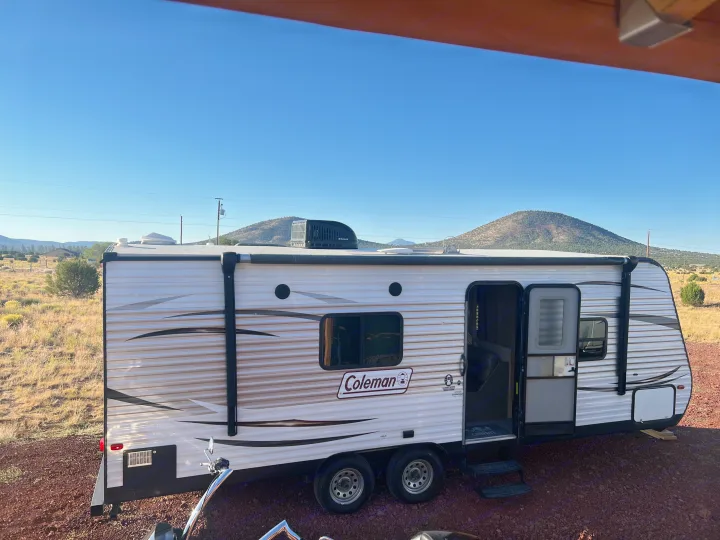 White & gray Coleman travel trailer rv rental with open door, parked on gravel in a sunny, arid landscape with hills.. Dutchmen Coleman Lantern LT 2016