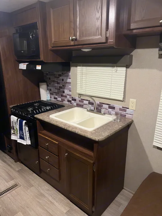 Functional RV rental kitchen with dark wood cabinets, stove, dual sink, microwave, and light wood floor.. Dutchmen Coleman Lantern LT 2016