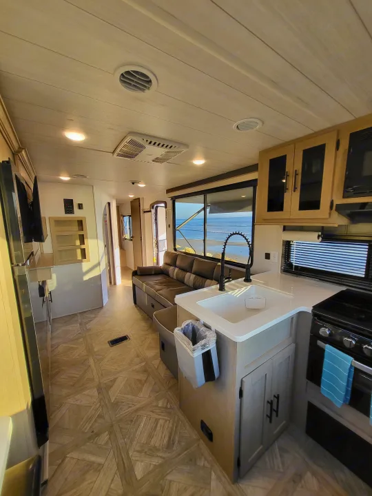 Modern RV rental interior with full kitchen, sofa, and a large window overlooking a sandy beach and ocean.. Forest River Wildwood 24 view 2024