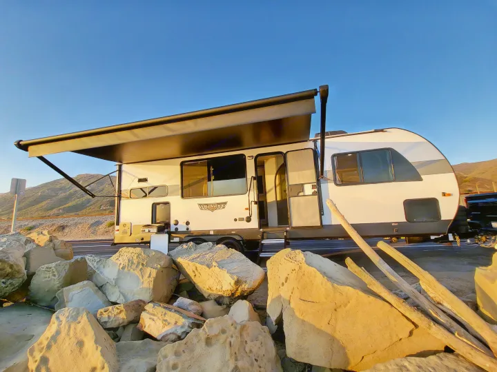 RV rental: A white travel trailer with awning extended, parked by coastal rocks & driftwood, with mountains under a clear sky.. Forest River Wildwood 24 view 2024
