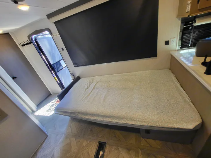 RV rental interior features a comfortable bed with patterned cover, a large window with black blind, and essential outlets.. Forest River Wildwood 24 view 2024
