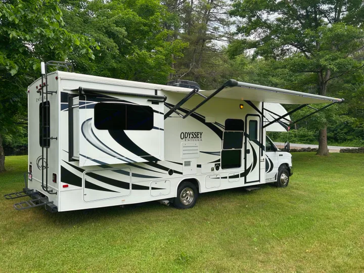 Odyssey Integra RV rental parked on green grass with trees. White motorhome with black graphics, extended awning, and rear ladder.. Entegra Coach Odyssey 2020