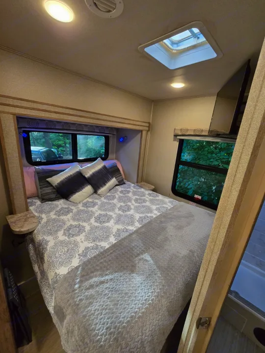 Bright RV rental bedroom: cozy bed with patterned duvet, skylight, windows showcasing lush trees, and small side tables.. Entegra Coach Odyssey 2020