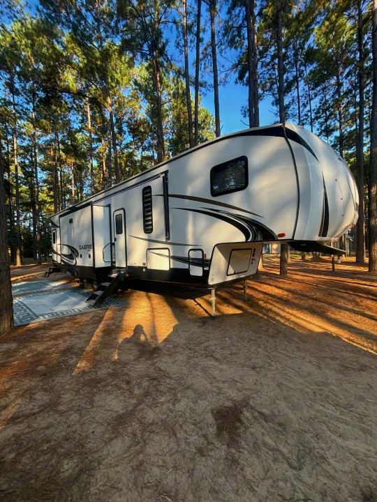 Spacious fifth-wheel rv rental parked at a serene pine forest campsite with outdoor rugs under a clear blue sky.. Forest River Sabre 2022