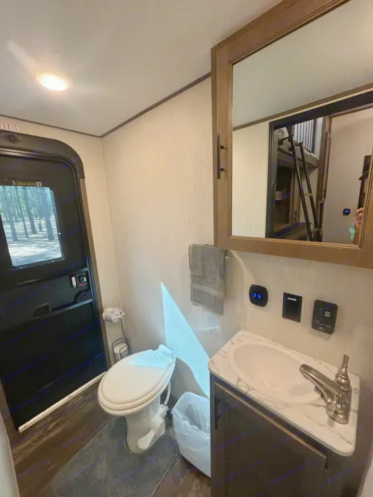 Compact RV rental bathroom with toilet, sink, and mirrored cabinet. View outside shows trees, reflection reveals bunk beds.. Forest River Sabre 2022
