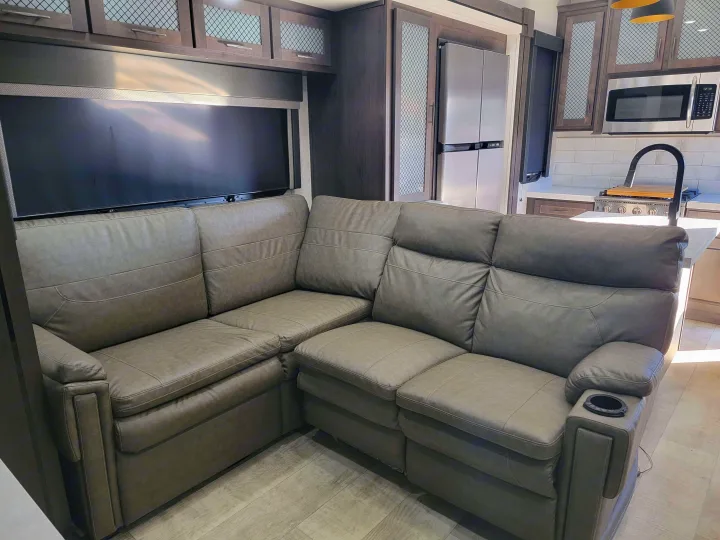 Modern RV rental interior: large L-shaped sofa with cup holders, window, and kitchen area featuring a microwave & refrigerator.. Grand Design Momentum 2022