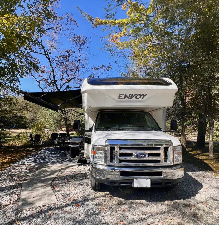 Experience nature with this white Envoy RV rental! Parked at a tree-lined campsite under a clear blue sky.. Jayco Envoy 100 2018