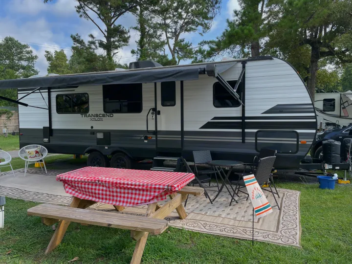 Relaxing RV rental site featuring a white travel trailer, shaded outdoor seating, picnic table with checkered cloth, and rug.. Grand Design Transcend Xplor 2021