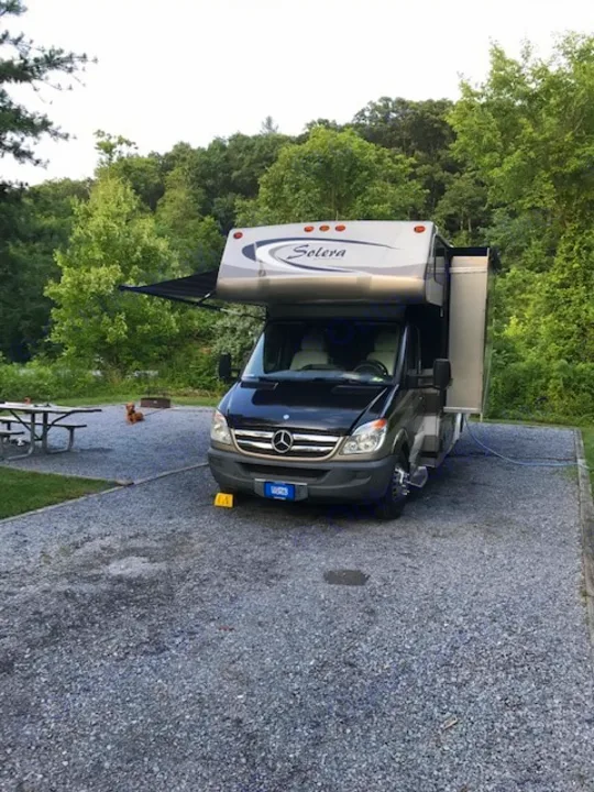 This Solera RV rental is parked on a gravel campsite with its awning extended, surrounded by trees. A small dog rests nearby.. Forest River Solera 24R 2014