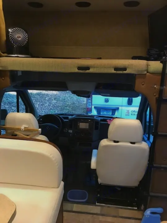 RV rental interior: Front cockpit view with white swivel seats, dashboard, and overhead bunk. A fan is on the bunk bed.. Forest River Solera 24R 2014