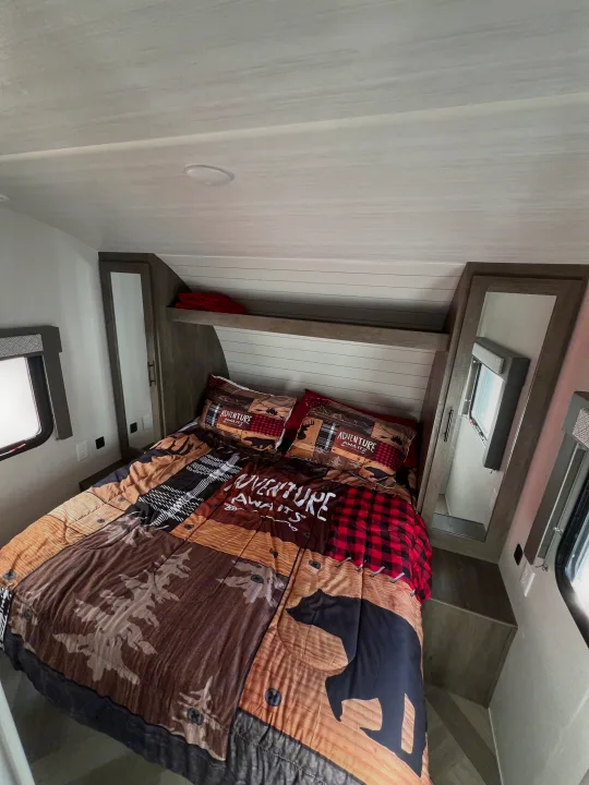 RV rental interior: A cozy bedroom featuring "Adventure Awaits" rustic bedding, windows, and compact storage.. Forest River Wildwood X-Lite 2020
