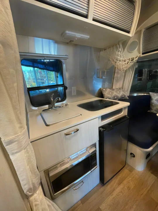 Inside this RV rental, a compact travel trailer kitchen: sink, stovetop, microwave, and fridge, with a cozy dinette.. Airstream Bambi 2022